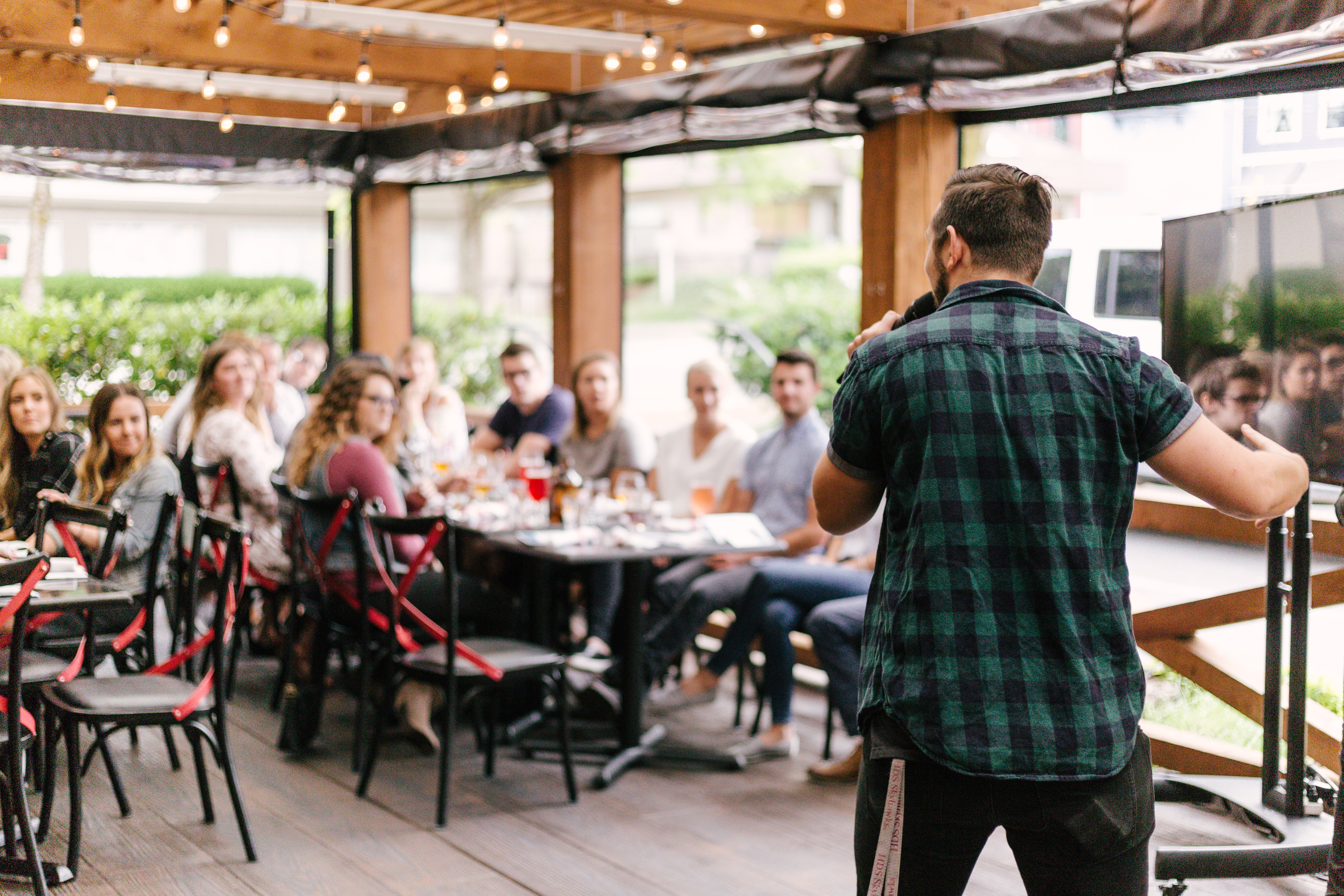 A man in his twenties in a green plaid shirt with short sleeves stands at the front of a room full of people. He is speaking into a microphone. It appears to be a restaurant, with chairs set up at long tables. There are several people in their twenties or thirties sitting at the tables, looking at the man speaking.
